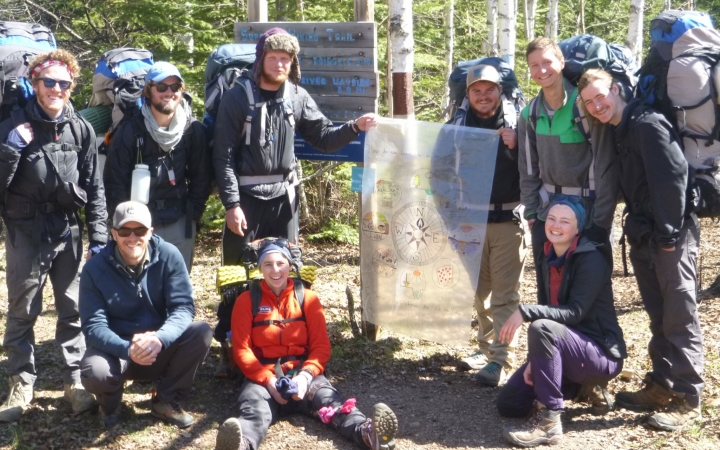 A group of students wearing backpacks pose for a photo with an Outward Bound sign. 